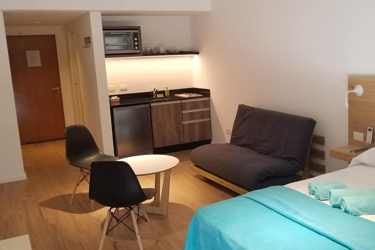 A hotel room with a bed, a desk, a chair, a kitchenette, and a door.