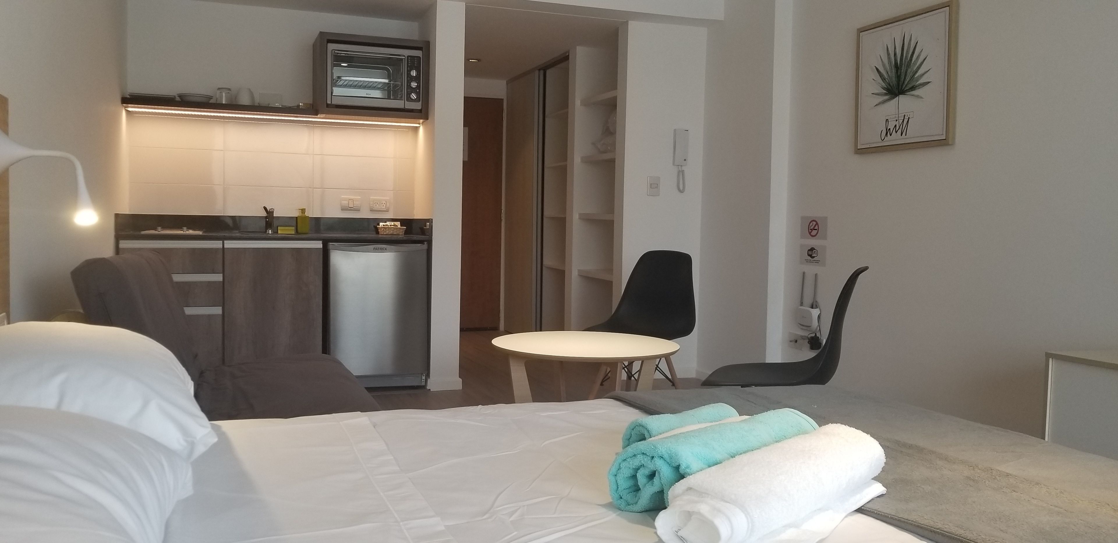 A hotel room with a bed, a desk, a chair, a small table, a kitchenette, and a bathroom.