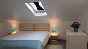 Une chambre avec un puits de lumière, un lit recouvert d’une couverture bleue, deux lampes de chevet, une petite commode et une plante en pot.