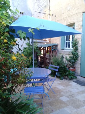 Mesa e cadeiras azuis ao ar livre, sob um guarda-sol azul, rodeadas por plantas em vasos e um muro de pedra.