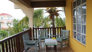 Ein Balkon mit Fliesenboden, Holzgeländer und zwei Stühlen mit Kissen. In der Mitte steht ein Tisch mit Tischdecke und Blumenvase.