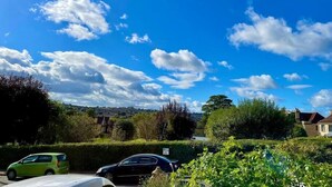 Un barrio residencial con cielo azul despejado y nubes dispersas. Hay dos coches aparcados en la calle, un coche verde y uno negro.
