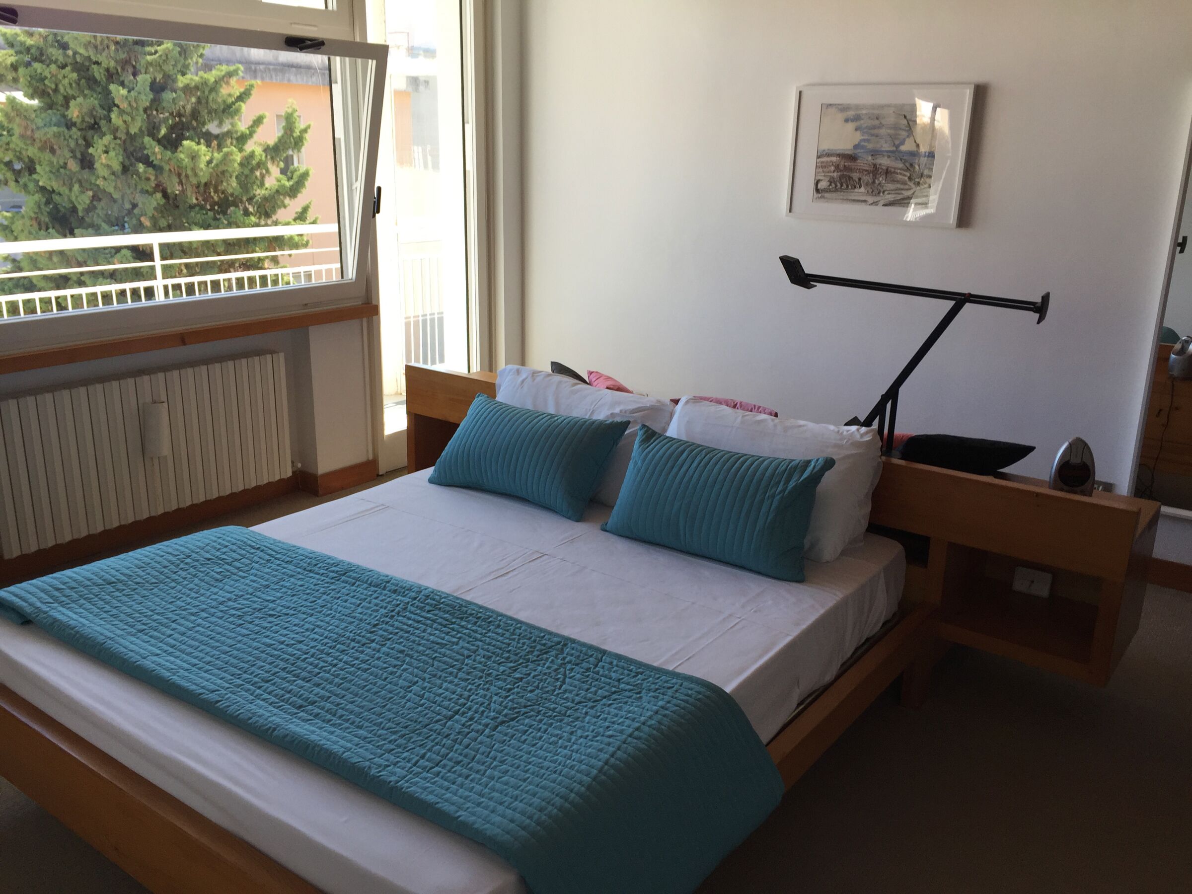 Un lit recouvert d’une courtepointe turquoise, avec du linge de lit blanc et deux oreillers turquoise. Une table de chevet avec une lampe et une petite horloge est visible. Un tableau encadré est accroché au mur.