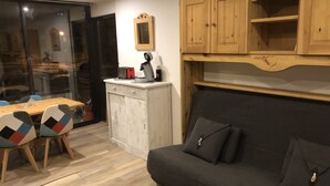 A modern living room with a grey sofa, wooden cabinets, a dining table with chairs, and a kitchen area.