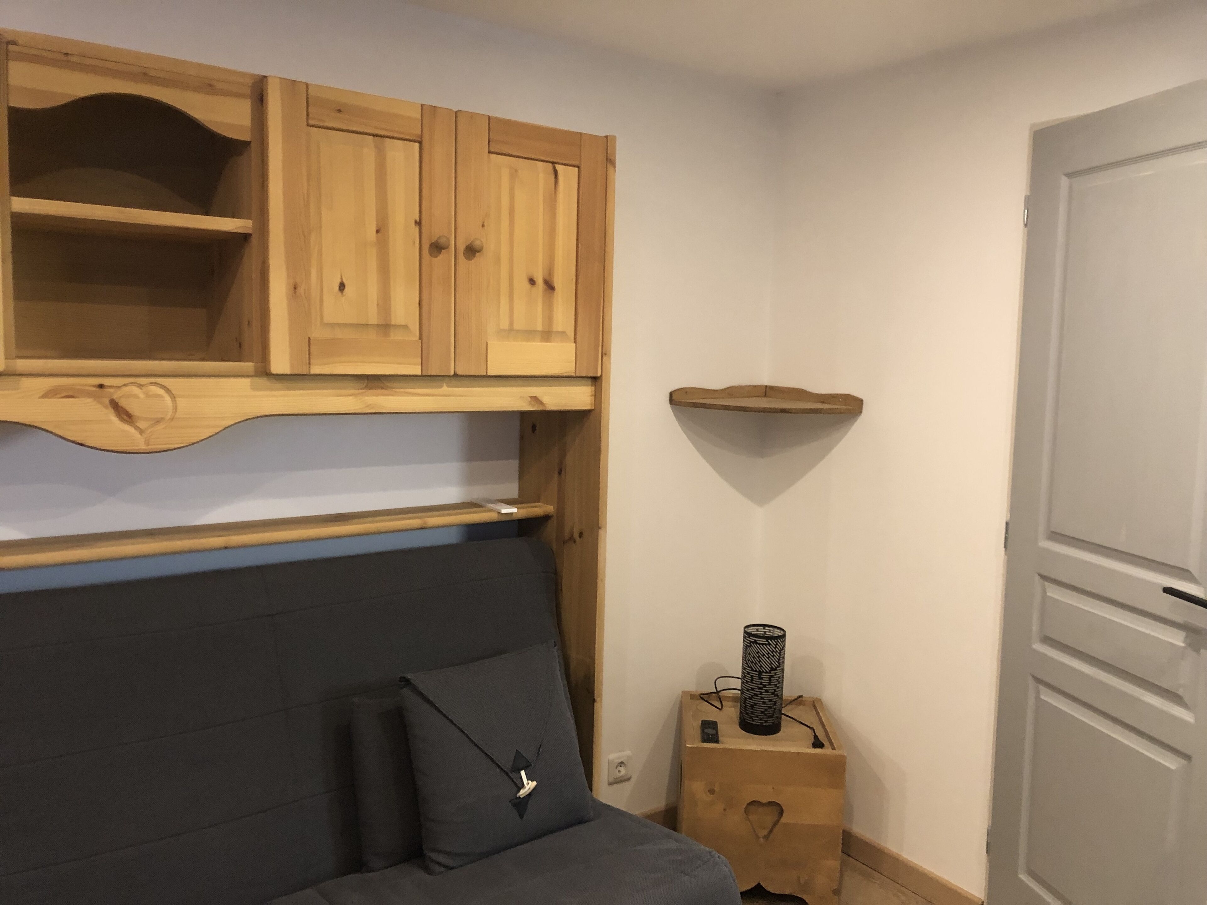 Una habitación con un sofá de color oscuro, un armario de madera con estantes, una mesita de madera con una lámpara y una puerta blanca.