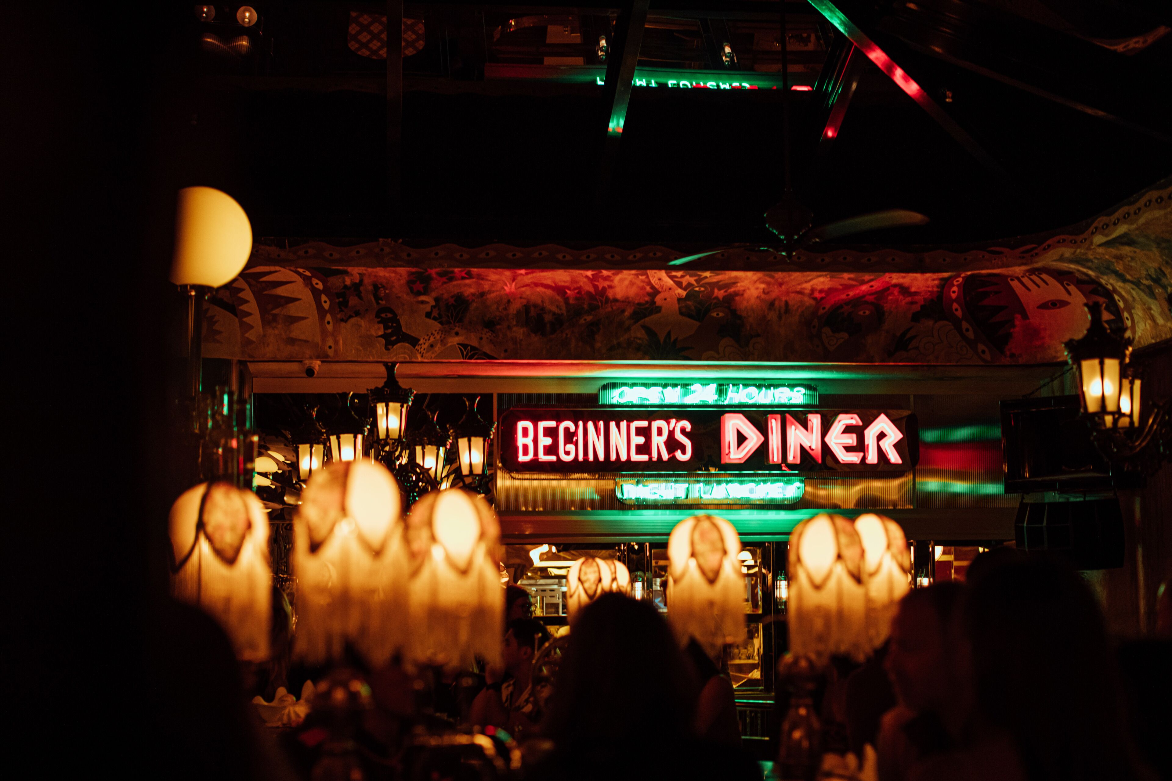 A dimly lit bar with a neon sign reading "BEGINNER'S DINER" and a patterned ceiling.