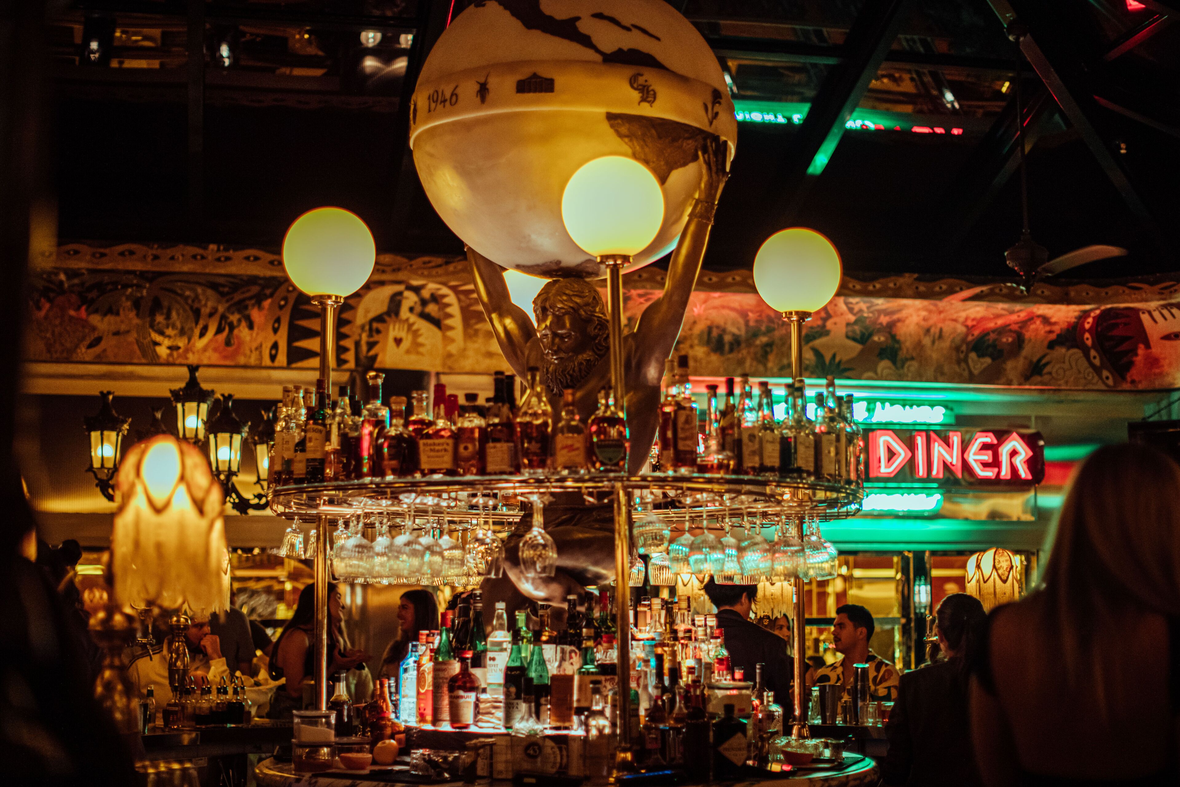 A bar with a globe centerpiece, various bottles, and a neon diner sign in the background.