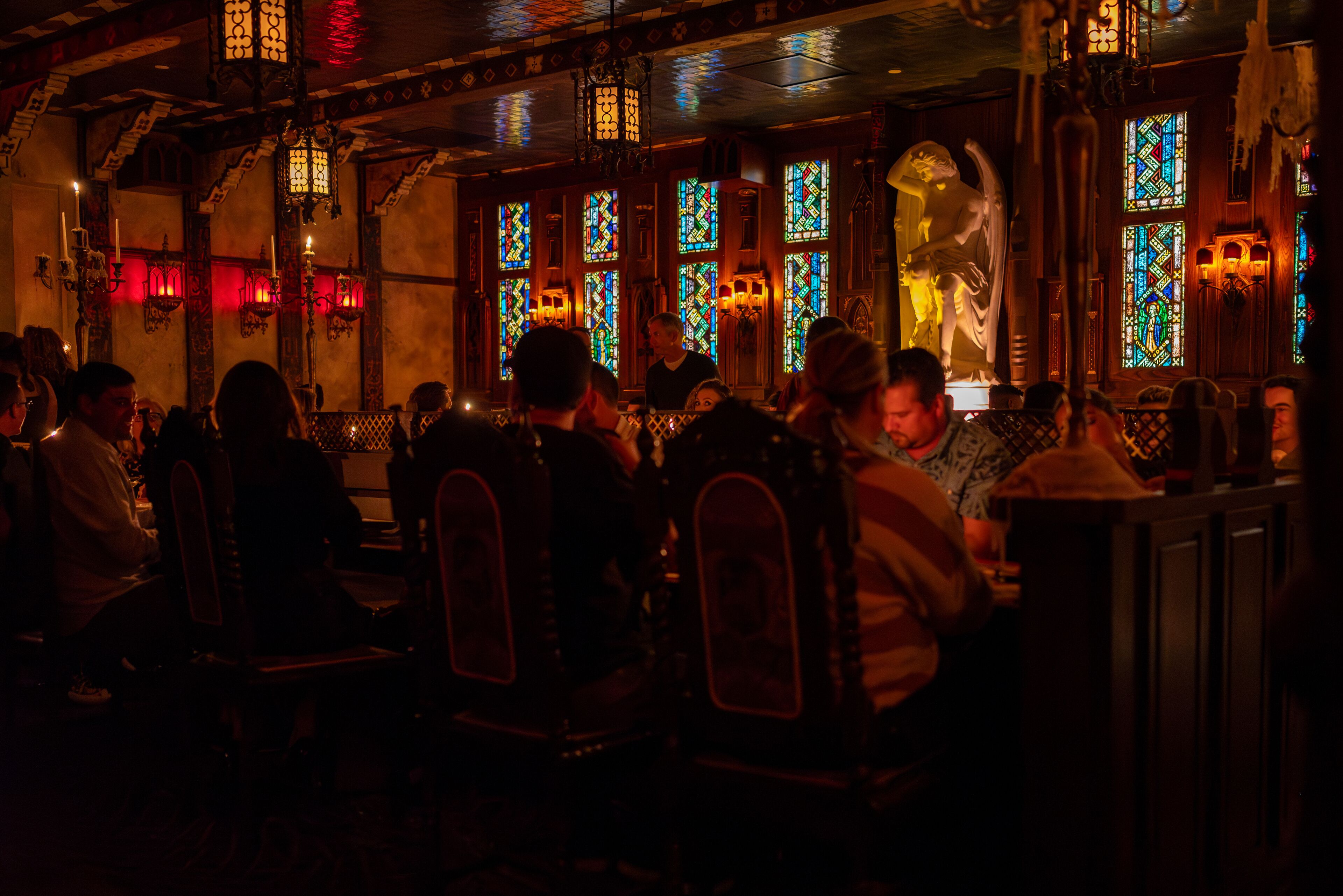 A dimly lit restaurant with patrons seated at tables, ornate stained glass windows, and chandeliers.