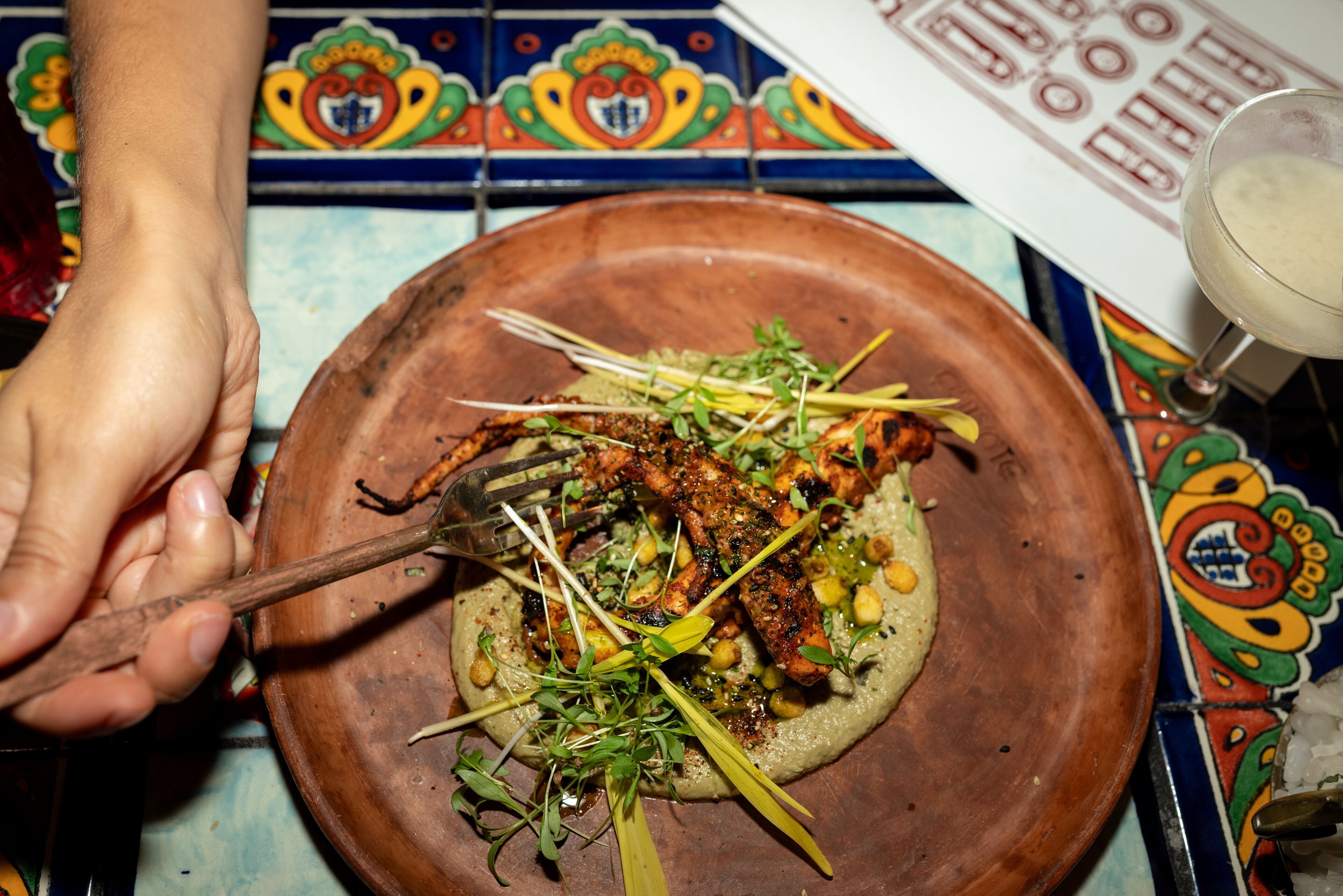 Une main tenant une fourchette au-dessus d’une assiette contenant des aliments grillés, agrémentés de légumes verts et de pois chiches, sur une table colorée recouverte de carreaux.