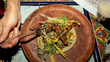 Une main tenant une fourchette au-dessus d’une assiette contenant des aliments grillés, agrémentés de légumes verts et de pois chiches, sur une table colorée recouverte de carreaux.