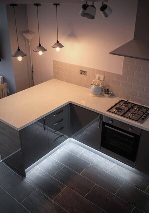 Une cuisine moderne équipée d’une cuisinière à gaz, d’une bouilloire et de luminaires suspendus.