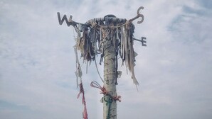 Un albero con una croce sulla cima, decorato con diversi oggetti, sullo sfondo di una spiaggia e dell'oceano.
