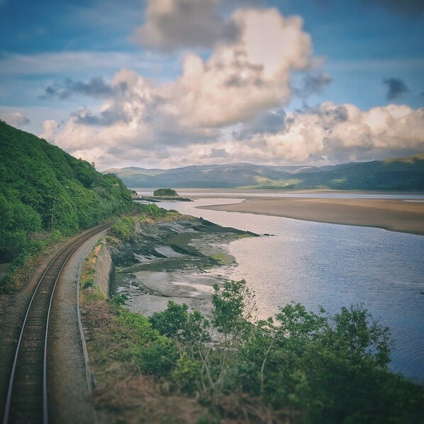 A railway track curving alongside a river with a view of distant hills and a partly cloudy sky.