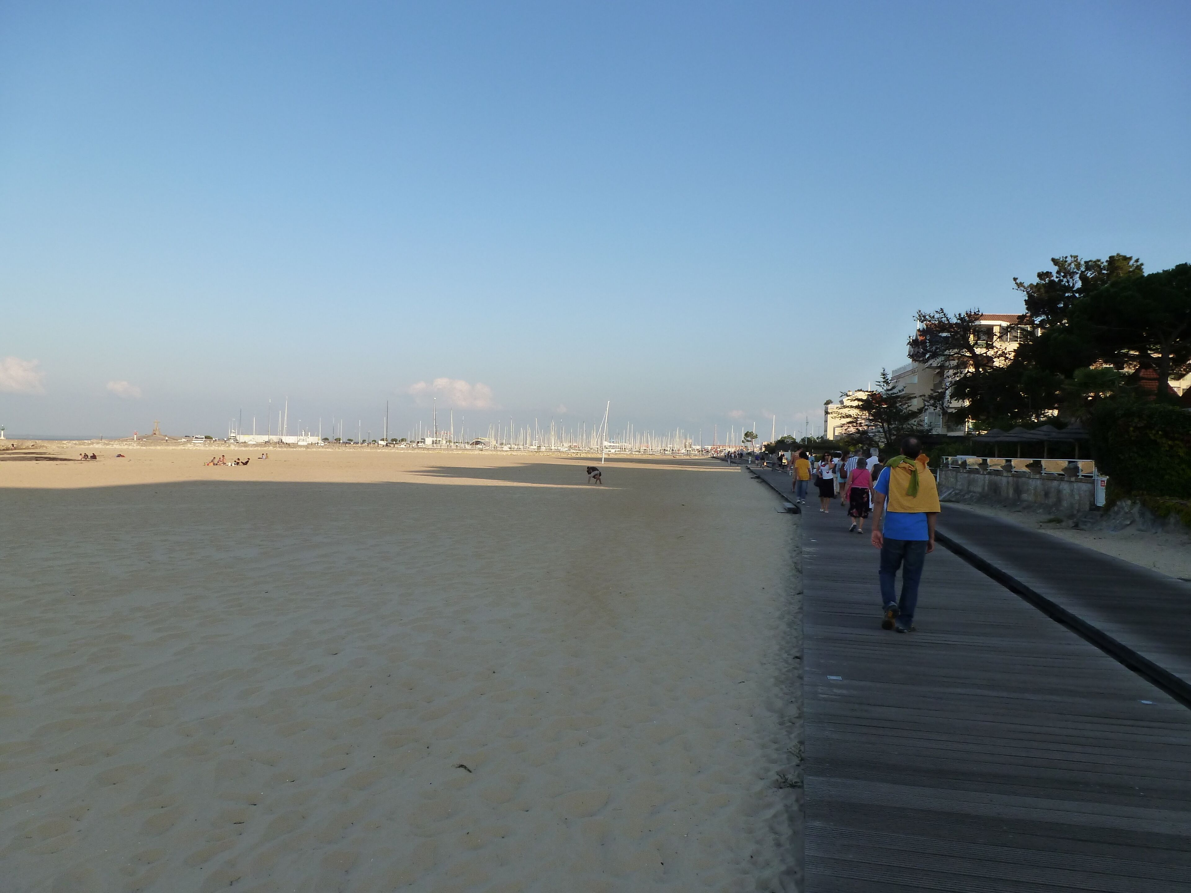 A beach with a wooden boardwalk, a sandy shore, and sailboats in the distance.