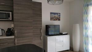 Een moderne keuken met een magnetron, waterkoker en koffiezetapparaat. Er is een flatscreen televisie op een wit kastje, een schilderij aan de muur en een gordijn met een patroon.