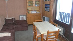 Ett mysigt vardagsrum med en brun soffa, ett matbord i trä med stolar, ett skåp med glasdörrar, en tv och ett fönster med utsikt över en balkong.