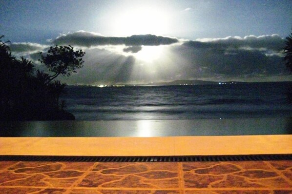 Une vue sur la piscine avec un coucher de soleil sur l’océan, un ciel dégagé et les lumières lointaines de la ville.