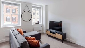 Una sala de estar moderna con un sofá gris, cojines naranjas, un mueble de televisión negro y ventanas amplias con vista a edificios.
