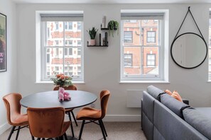 A dining area with a round table, four chairs, a sofa, and two windows with a view of buildings.