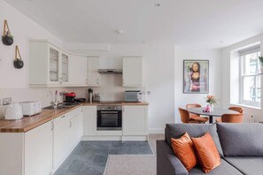 A modern kitchen with white cabinets, a microwave, and a toaster. There is a dining area with a table and chairs, and a gray sofa with orange cushions.