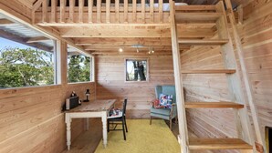 Interior de una cabaña de madera con cama en altillo, escritorio, silla y ventana con vista a follaje.