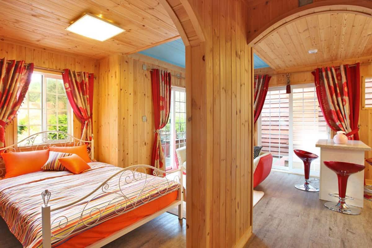 Una cabaña de madera con una cama, cortinas rojas y una pequeña zona de bar con sillas rojas.