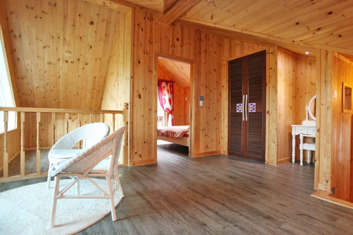 Interior de una cabaña de madera con una cama, una silla de mimbre blanca, un aparador y un espejo.