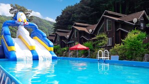 Ein Swimmingpool mit einer blauen aufblasbaren Rutsche, umgeben von Holzhäusern und Grünpflanzen.