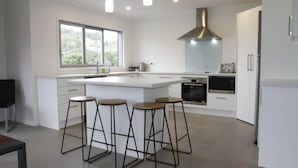A modern kitchen with a central island, bar stools, a large window, and built-in appliances. - CooksEstuaryEscape-Waterfront at your doorstep (Price enquire for small groups) (Cooks Beach)