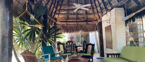 A thatched-roof lounge area with wooden furniture, including chairs and a sofa, and a ceiling fan.