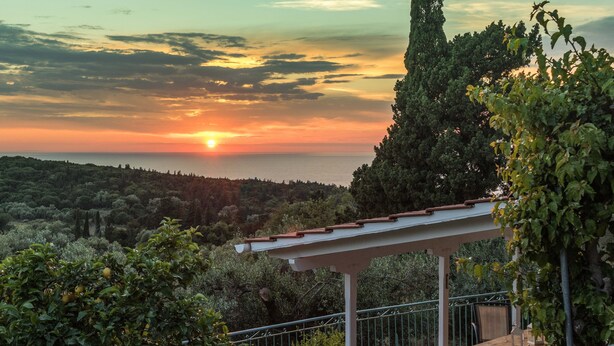 A terrace with a view of the sunset over the sea, surrounded by greenery and potted plants. - Stylish Stone Cottage with Pool near Fiskardo (Agrilias)