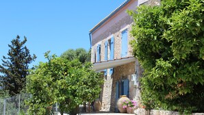 A stone building with blue shutters, a garden with trees and a large vase of flowers, and a clear blue sky. - Stylish Stone Cottage with Pool near Fiskardo (Agrilias)