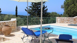 A pool area with two blue lounge chairs, a white umbrella, and a stone wall. - Stylish Stone Cottage with Pool near Fiskardo (Agrilias)
