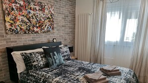 Une chambre à coucher moderne avec une couvre-lit à motifs noir et blanc, un tableau représentant un paysage urbain au-dessus du lit, un mur en briques et une fenêtre ornée de rideaux légers.
