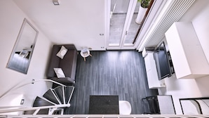 A modern interior with a grey tiled floor, a black sofa with cushions, a white chair, a small table with a magazine, a white radiator, and a TV on a white cabinet.