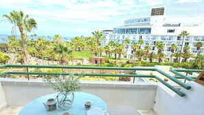 A balcony with a table set for two, a vase, and a view of a hotel building and palm trees.