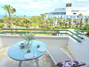 A balcony with a table set for two, a vase, and a view of a hotel building and palm trees.