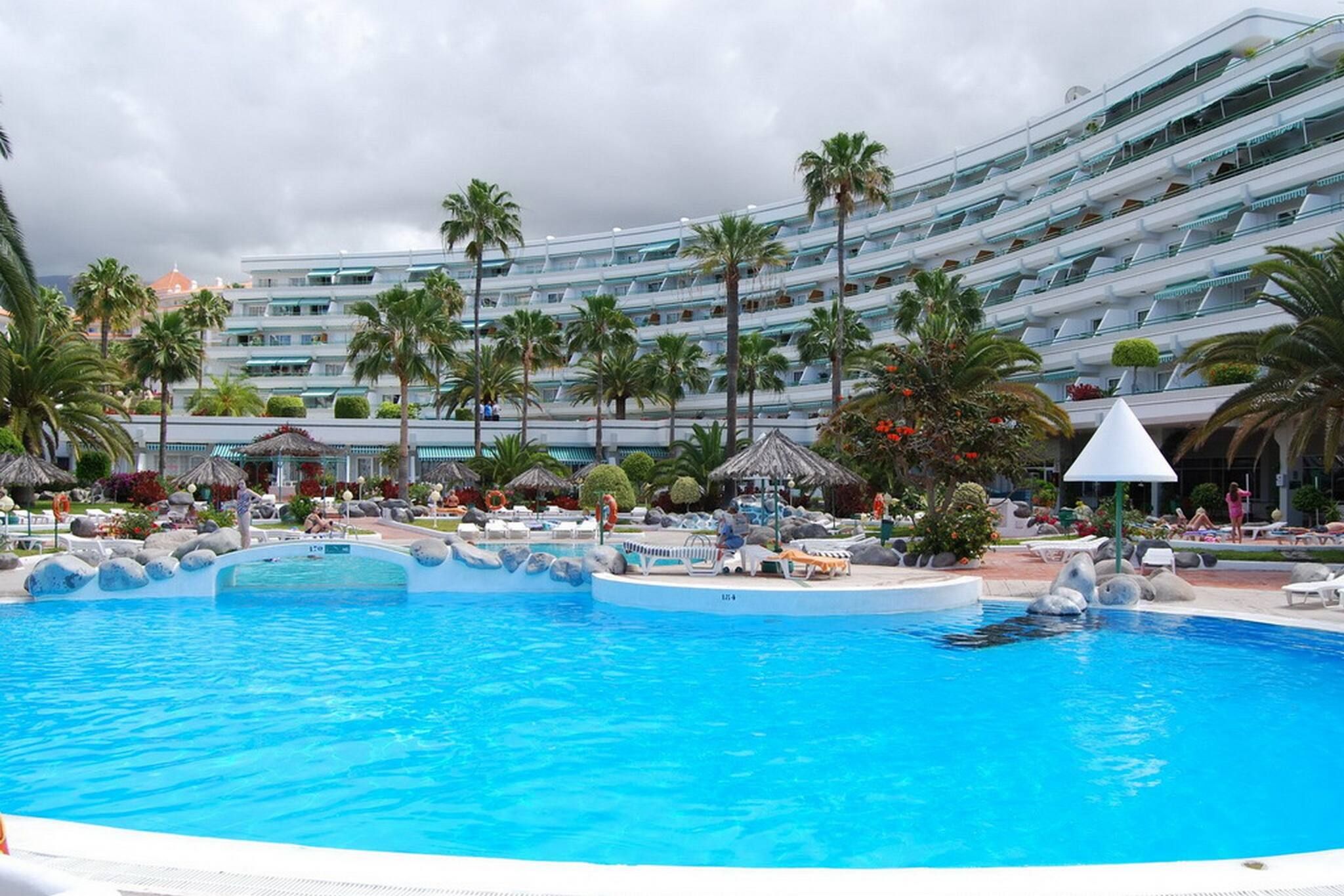 A large swimming pool with lounge chairs and palm trees, surrounded by a multi-story hotel building.