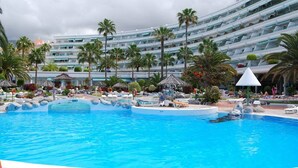 A large swimming pool with lounge chairs and palm trees, surrounded by a multi-story hotel building.