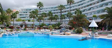 A large swimming pool with lounge chairs and palm trees, surrounded by a multi-story hotel building.