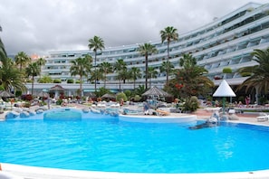 A large swimming pool with lounge chairs and palm trees, surrounded by a multi-story hotel building.