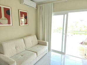 Een woonkamer met een beige bank, twee ingelijste abstracte kunstwerken, een schuifdeur naar een buitenruimte en een airconditioner aan de muur bevestigd.