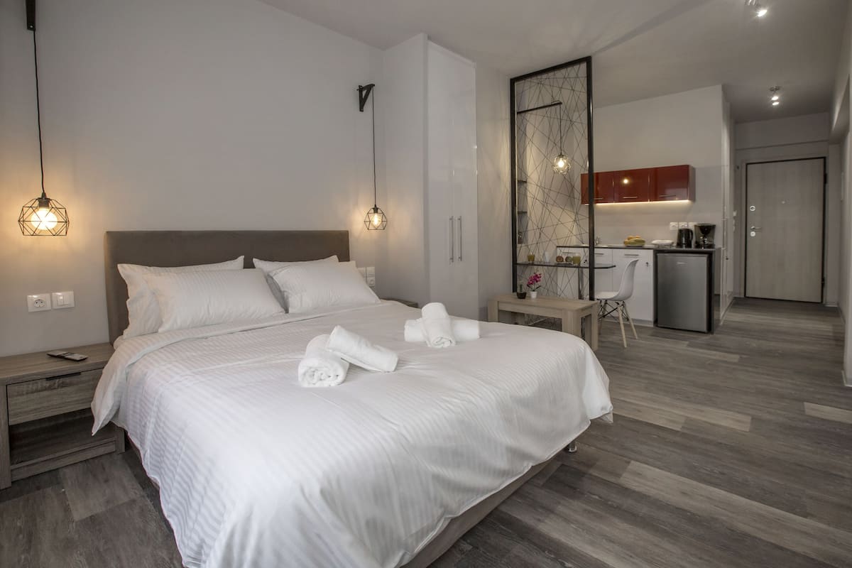 A modern hotel room with a large bed, two bedside lamps, a wooden nightstand, a desk with a chair, and a kitchenette in the background.