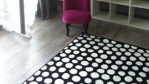 A room with a grey wall, a white shelf unit, a pink chair, a black and white polka dot rug, and a window with curtains.