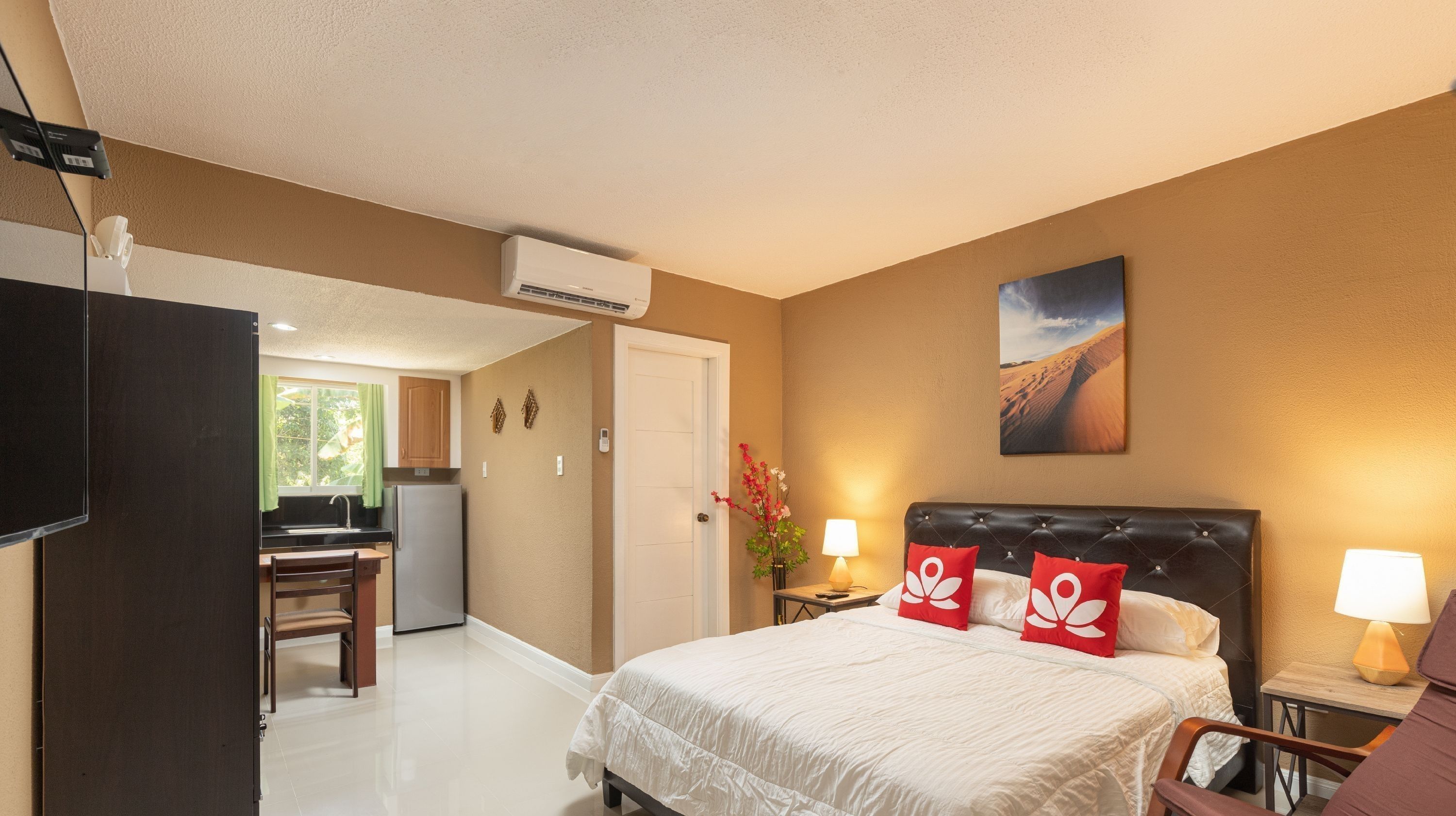 A hotel room with a bed, bedside lamps, a painting, a desk, and a kitchenette.