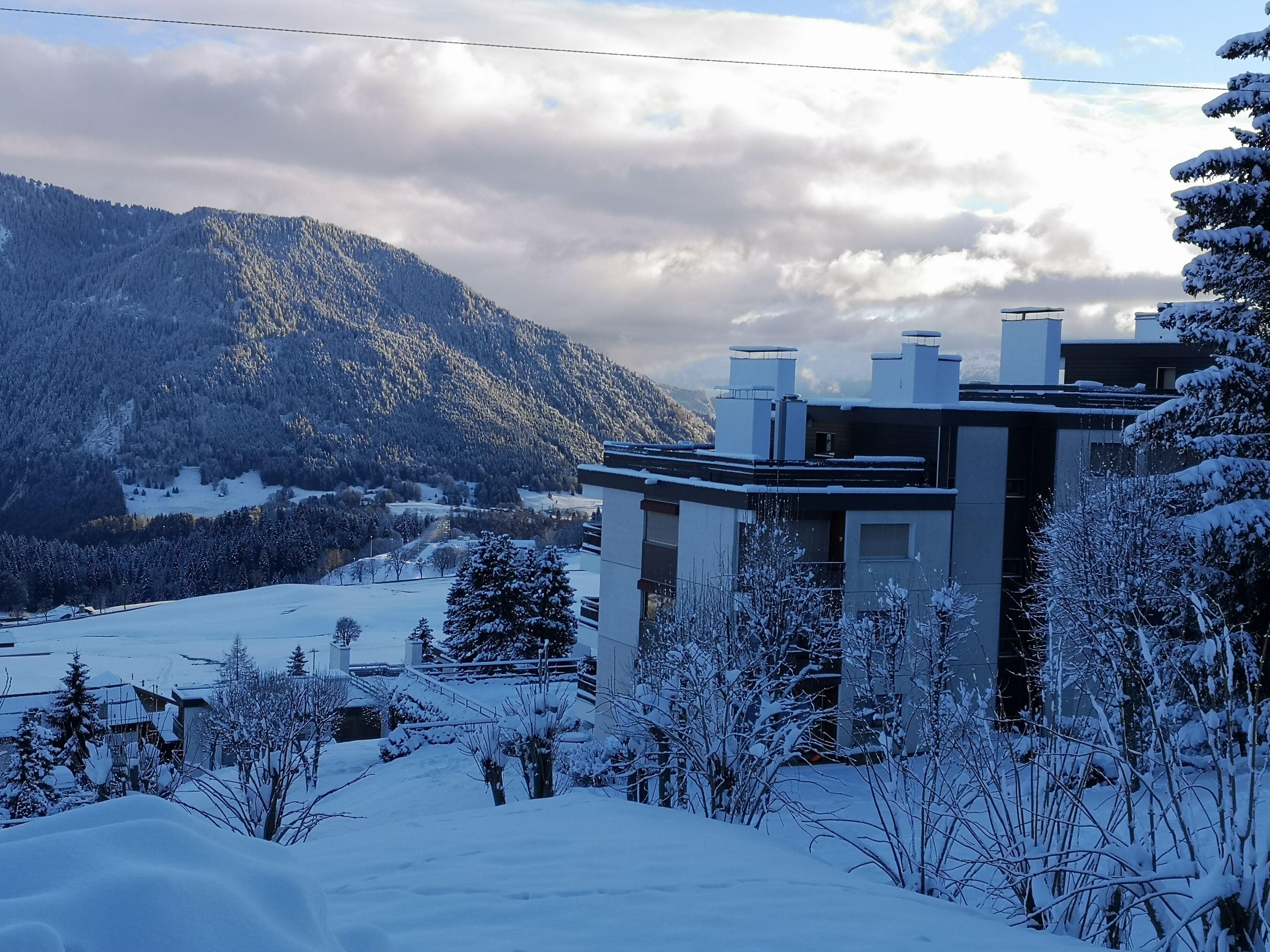 A modern building with multiple chimneys, surrounded by snow-covered trees and a mountainous backdrop.