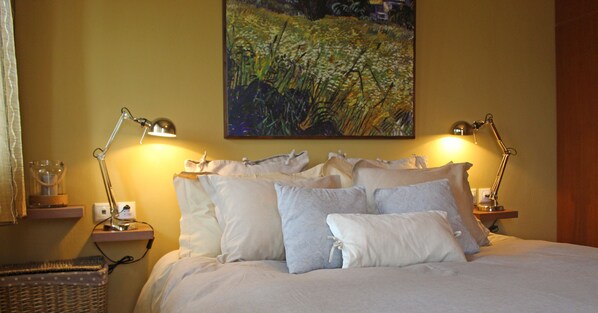 A neatly made bed with white linens and multiple pillows, two bedside lamps, a painting of a field on the wall, and a small wooden nightstand with a lamp.