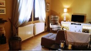 A living room with a sofa, armchair, and a television. There are two radiators, a lamp, and a cabinet with bottles.