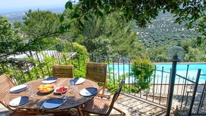 A wooden table set with plates, cutlery, and glasses, overlooking a swimming pool and lush greenery.