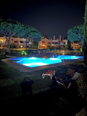 Una piscina nocturna con sillones de descanso y un muro de piedra.